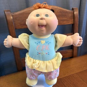May include: A Cabbage Patch Kid doll with a peach-colored face, green eyes, and orange yarn hair. The doll wears a light blue top with "CPK" embroidered on it, yellow sleeves, and floral patterned pants. It stands on a white base.