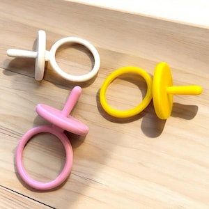 May include: Three colorful, ring-shaped toys in white, pink, and yellow. Each toy has a circular ring and a small, perpendicular post. The toys are arranged on a light-colored wooden surface.