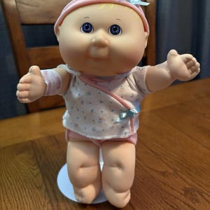Puede incluir: Muñeca Cabbage Patch Kid vintage con ojos azules, que lleva un gorro rosa, una camisa blanca con un estampado pastel y pantalones cortos rosas. La muñeca tiene una venda en el brazo y está de pie sobre una base blanca.