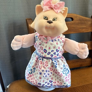 May include: A plush cat doll with a light peach complexion, green eyes, and freckles. The doll wears a pink hair bow, a pink collar with a flower and small bell, and a white dress featuring a colourful leopard print pattern in pink, blue, and grey.