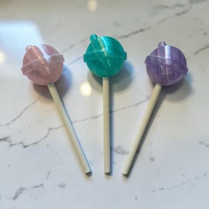 May include: Three colorful lollipops with white sticks. The lollipops are in shades of pink, teal, and purple. The candy heads have a textured design. The lollipops are arranged on a white marble surface.