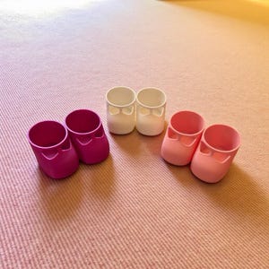 Puede incluir: Cuatro pares de zapatos de bebé en fucsia, blanco y rosa claro. Los zapatos son de material sólido y tienen un estilo Mary Jane con punta redonda y una correa en la parte superior. Los zapatos están colocados sobre una alfombra rosa.