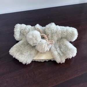 May include: A light sage green faux fur bolero jacket featuring long sleeves and a round neckline. The front closure consists of two matching faux fur pom-poms attached to a braided beige cord tie, providing a soft and textured appearance.