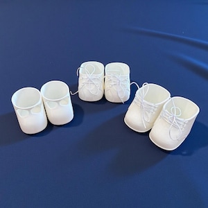 May include: Three pairs of white doll shoes made of molded plastic. One pair is plain with cutouts, while the other two pairs feature molded lace details and thin white string laces tied in bows across the top of the footwear.