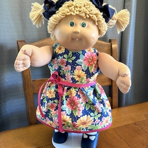 Puede incluir: Muñeca vintage con cabello rubio de hilo peinado en coletas, adornada con lazos azul marino. La muñeca viste un vestido floral azul con una cinta rosa y zapatos a juego. La muñeca está sobre una base blanca.