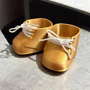 Può includere: Un paio di fioriere dorate a forma di scarpa, stampate in 3D. Ogni fioriera ha lacci bianchi e un design classico della scarpa. Le fioriere sono appoggiate su una superficie grigia.