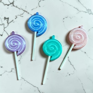 Pode incluir: Quatro pirulitos coloridos em espiral nas cores azul, roxo, verde-azulado e rosa com palitos brancos. Os pirulitos estão dispostos sobre uma superfície de mármore branco e cinza. Os pirulitos têm um acabamento brilhante.