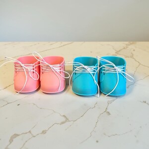 May include: Two pairs of small, smooth, matte-finish shoes, one pair in light pink and one pair in bright turquoise. Each shoe features a rounded toe, a low-cut ankle opening, and thin white laces threaded through eyelets on the sides.