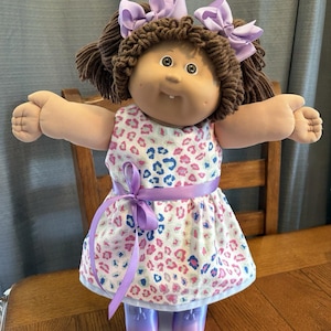 May include: A doll with brown yarn hair styled in pigtails with two light purple bows. The doll has a tan complexion, green eyes, and a single tooth. It wears a white dress with a pink and blue leopard print, a purple ribbon waist tie, and purple shoes.