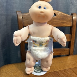 May include: A vintage Cabbage Patch Kids doll, featuring a soft, flesh-toned face with blue eyes and a tuft of blonde hair. The doll is wearing a white diaper with a colorful print. It is standing on a round base.