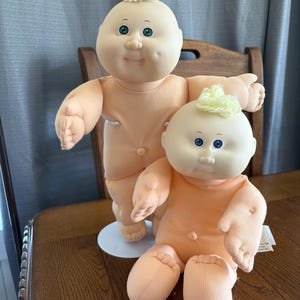 Op de afbeelding: Twee poppen met zachte stoffen lichamen en ronde hoofden. De grotere pop heeft groene ogen en een kleine pluk geel garen als haar. De kleinere pop heeft blauwe ogen en een soortgelijke gele pluk. Beide poppen hebben een perzikkleurige huid.