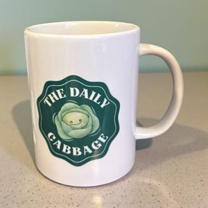 May include: White ceramic mug with a green and white graphic of a smiling cabbage and the words "THE DAILY CABBAGE" in a dark green border. The mug has a curved handle.