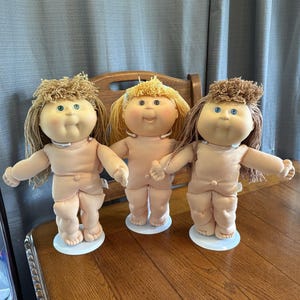 Könnte beinhalten: Drei Vintage Cabbage Patch Kids Puppen mit heller Haut, blauen Augen und verschiedenen Haarfarben. Jede Puppe steht auf einem weißen Sockel, die Arme ausgestreckt. Die Puppen werden auf einer Holzoberfläche präsentiert.