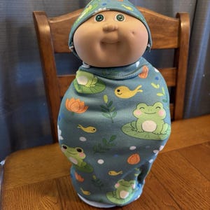 May include: A doll wrapped in a teal swaddle blanket and matching hat. The blanket features a pattern of frogs on lily pads, yellow fish, and orange flowers. The doll has a round face, rosy cheeks, and green eyes.