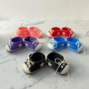 May include: A collection of five pairs of miniature sneaker-style shoes in pink, red, purple, blue, and black. Each shoe features a white toe cap, white laces, and a white star emblem on the side. The shoes are arranged in a group on a white surface.