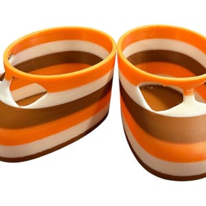 May include: A pair of small, striped shoes in shades of orange, white, and brown. The shoes have a rounded toe and a strap across the top. The shoes are likely for a baby or small child.
