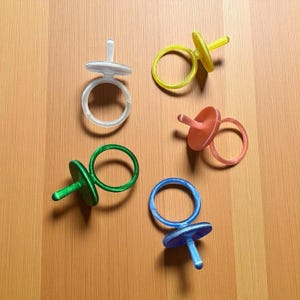 Może przedstawiać: Pięć kolorowych pierścieni w kształcie smoczka, w kolorze białym, żółtym, koralowym, zielonym i niebieskim. Pierścienie wykonane są z półprzezroczystego materiału i ułożone na jasnobrązowej drewnianej powierzchni.