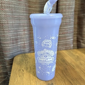 Op de afbeelding: Een lichtpaarse plastic beker met een bijpassend deksel en een flexibel rietje. De beker is voorzien van witte bedrukte afbeeldingen, waaronder het Cabbage Patch Kids-logo, de tekst Babyland General Hospital en het jaartal 2004, omringd door kleine krullen.
