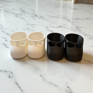 May include: Four small, cylindrical containers. Two are white, and two are black. Each has a cutout design near the top, resembling a bow tie. The containers are arranged in a row on a marble surface.