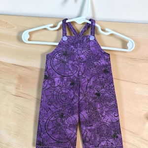 Op de afbeelding: Een paarse tuinbroek met een patroon van zwarte spinnenwebben en spinnen. Het kledingstuk heeft twee schouderbanden die aan de voorzijde zijn bevestigd met lichtpaarse ronde knopen, hangend aan een witte plastic hanger tegen een effen achtergrond.