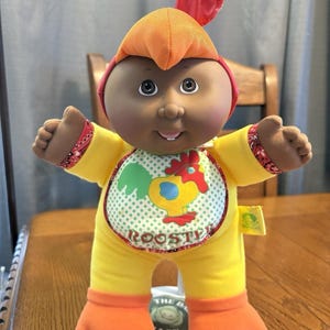 May include: A Cabbage Patch Kids doll dressed as a rooster. The doll has a dark complexion, wearing a yellow jumpsuit with orange feet and a matching cap. A bib features a rooster graphic and the word "ROOSTER".