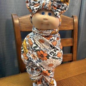Può includere: Bambola Cabbage Patch Kid avvolta in un tessuto a stampa floreale. La bambola ha gli occhi blu e un'espressione neutra. Il tessuto presenta un motivo di fiori arancioni, gialli e bianchi. Un fiocco abbinato adorna la testa della bambola.