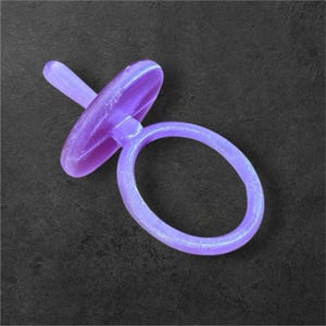 May include: A purple pacifier with a round handle and a flat, oval-shaped shield. The pacifier is made of a translucent material and is set against a dark background. The design is simple and modern.