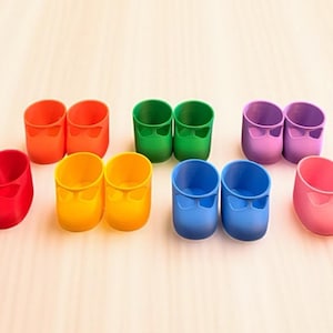May include: A collection of colorful, miniature shoes in various shades, including red, orange, yellow, green, blue, purple, and pink. The shoes have a rounded toe and a small bow detail. They are arranged in pairs on a light-colored surface.