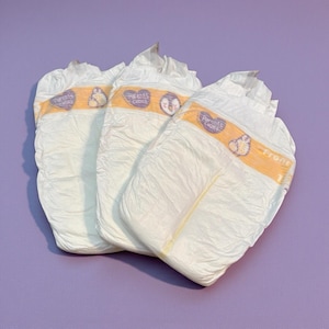May include: Three white baby diapers with yellow and orange accents. The diapers have a printed design and the words "Parents Choice" are visible. The diapers are arranged on a purple surface.