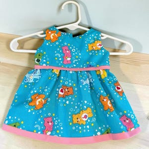 May include: A sleeveless dress in a bright turquoise blue fabric featuring a repeating pattern of colourful Care Bears, small yellow stars, and white hearts. The dress has a pink horizontal band at the waist and a matching pink hem at the bottom edge.