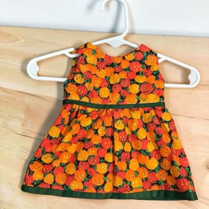 Op de afbeelding: Een mouwloze jurk met een all-over print van oranje en gele pompoenen op een zwarte achtergrond. De jurk heeft een donkergroene horizontale band in de taille en een bijpassende donkergroene bies langs de onderste zoom.