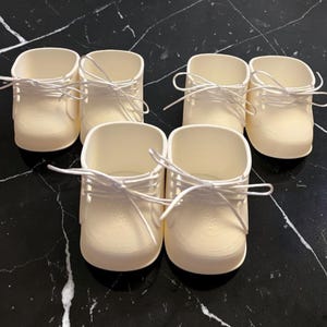 May include: A set of four off-white doll shoes, each featuring a rounded toe, a high-top ankle design, and thin white laces tied in a bow at the front. The shoes are constructed from a smooth, solid material with a moulded texture.