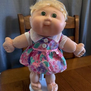 Peut inclure: Une poupée Cabbage Patch Kids vintage avec des cheveux blonds et des yeux verts. La poupée porte une robe à imprimé floral avec un col blanc et une bordure rose. La poupée est sur un support blanc.
