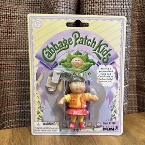 Peut inclure: Un porte-clés Cabbage Patch Kids représentant une petite poupée aux cheveux foncés, portant un gilet orange sur un haut jaune et une jupe rose, emballé sur une carte violette et blanche avec le texte Cabbage Patch Kids et Item #1198 Basic Fun.