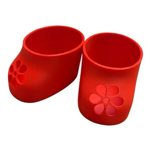 May include: Two bright red, open-topped containers. Each has a flower-shaped cutout on the side. The containers are cylindrical with a rounded base, resembling small shoes or cups. The background is plain white.