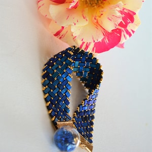 Op de afbeelding: Een handgemaakte blauwe kralenarmband met gouden accenten en een kleine blauwe glazen bol. De armband wordt getoond met een veelkleurige roos met roze en gele bloemblaadjes. Sieraden.