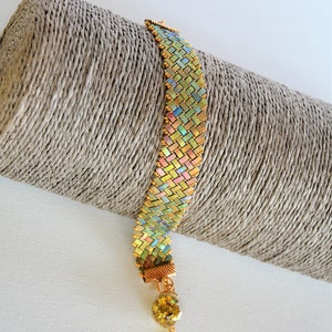Op de afbeelding: Een gouden en iriserende kralen armband met een gouden bedel en een gouden ketting. De armband ligt op een bruine achtergrond.