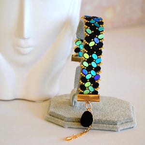 May include: A black beaded bracelet with a pattern of blue, green, and gold oval beads. The bracelet is displayed on a grey stand, with a black teardrop charm suspended from a gold chain.