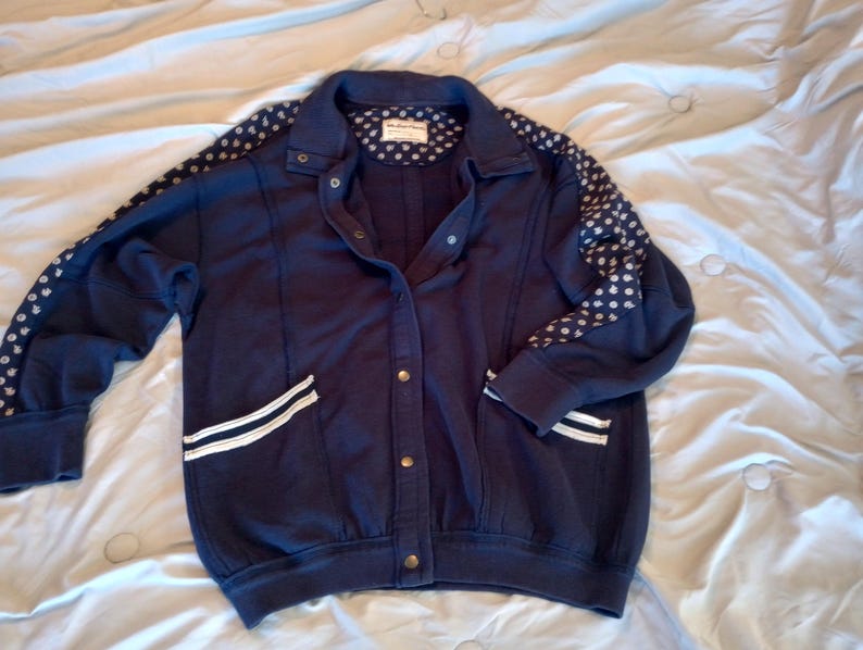 May include: Navy blue jacket with white polka dots on the sleeves and a white stripe detail on the pockets. The jacket has a button-up front and a collared neckline. The jacket is made of a soft material.