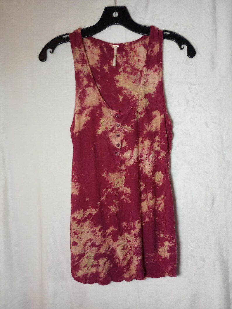 May include: A burgundy and beige tie-dye sleeveless top with a scoop neck and button-up placket, hanging on a black hanger. The fabric appears to be a soft, lightweight material. The top is a casual style.