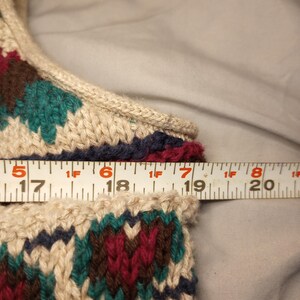 May include: Close-up of a beige knitted sweater with a colorful geometric pattern in shades of red, green, brown, and blue. A measuring tape is placed across the top of the sweater, showing the measurement of 17 inches.