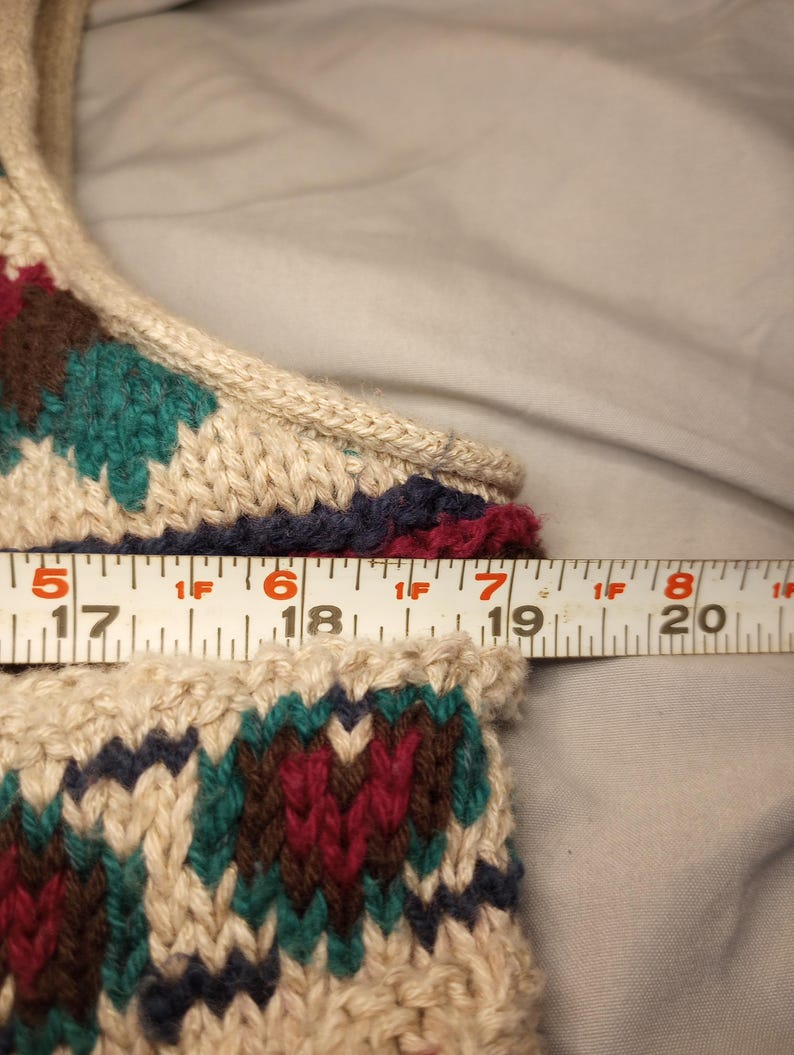May include: Close-up of a beige knitted sweater with a colorful geometric pattern in shades of red, green, brown, and blue. A measuring tape is placed across the top of the sweater, showing the measurement of 17 inches.