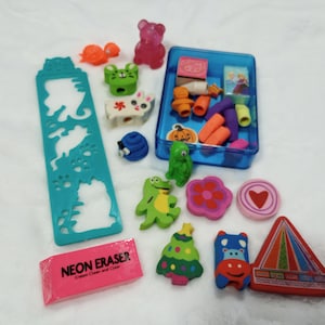 May include: A selection of colourful erasers in diverse shapes, such as animals, flowers, and geometric designs. A pink neon eraser is also present, along with a blue plastic container filled with more erasers. The erasers are scattered on a white surface.