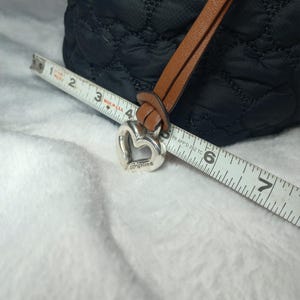 May include: Black quilted tote bag with a brown leather strap and a silver heart-shaped charm. The charm is engraved with the brand name "Brighton". A measuring tape is placed across the bag, showing measurements in inches.