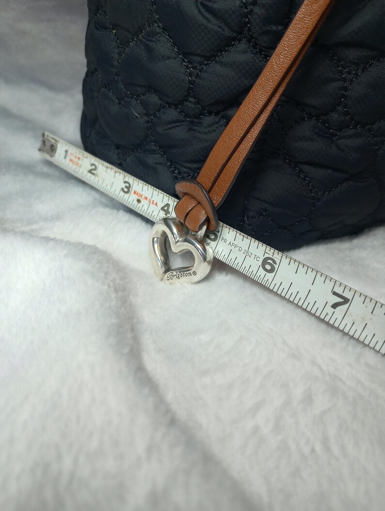 May include: Black quilted tote bag with a brown leather strap and a silver heart-shaped charm. The charm is engraved with the brand name "Brighton". A measuring tape is placed across the bag, showing measurements in inches.