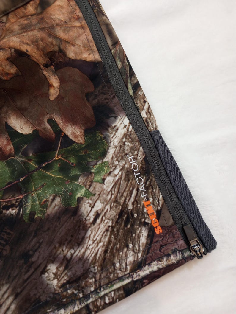 Puede incluir: Primer plano de una tela de camuflaje con una cremallera gris oscura. La tela presenta un patr&oacute;n de hojas y corteza marrones, verdes y beiges. El texto "SCENT-FACTOR" est&aacute; impreso en naranja.