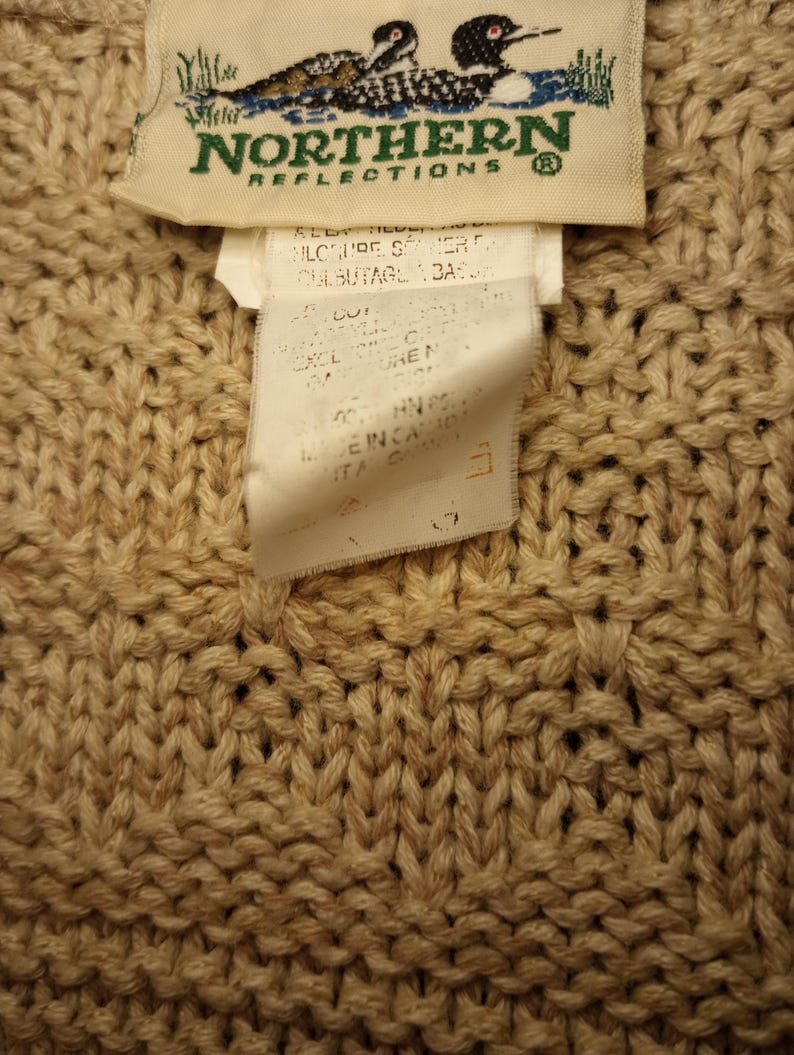 May include: A beige knitted sweater with a brown label that says "Northern Reflections" and features a pair of loons. The label also includes care instructions.