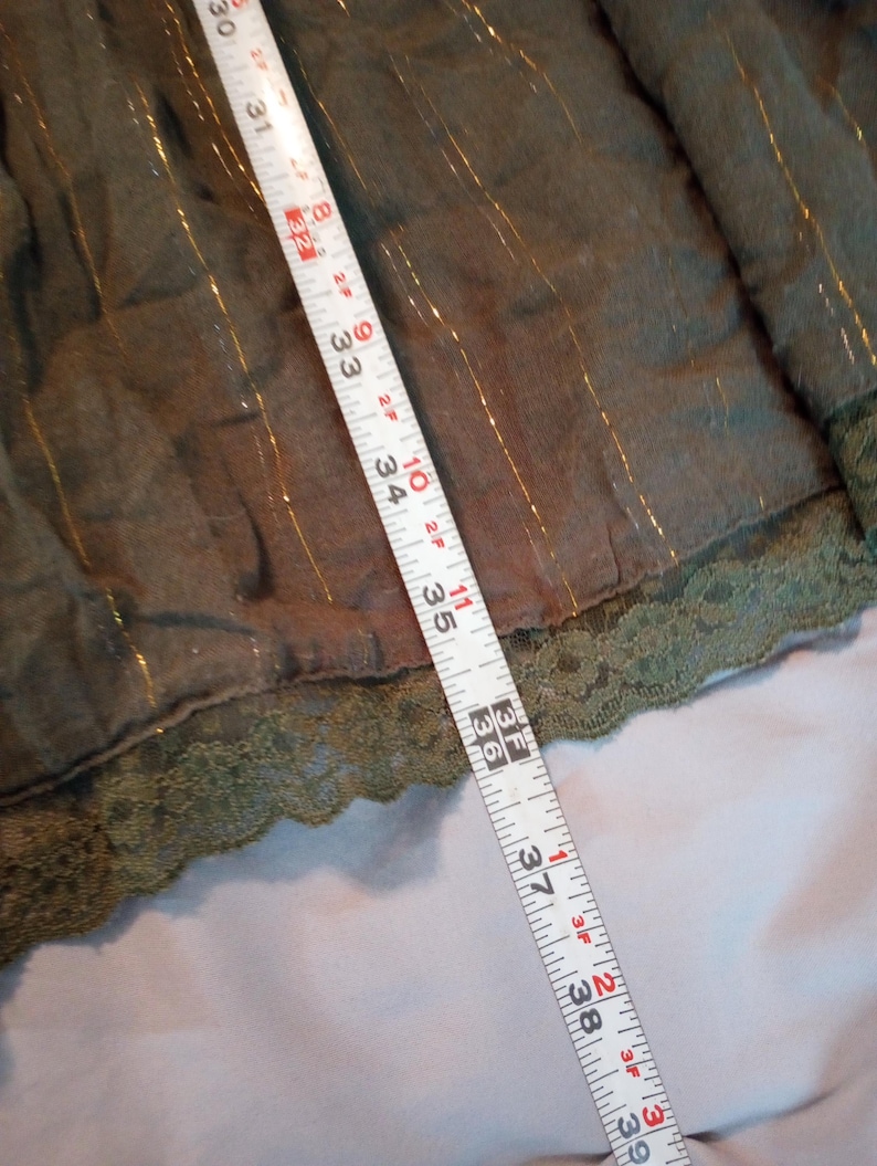 K&ouml;nnte beinhalten: Olivgr&uuml;ner Stoff mit goldenen Metallic-Streifen und einer Spitzenborte. Ein Ma&szlig;band zeigt, dass der Stoff etwa 76 bis 99 cm lang ist.