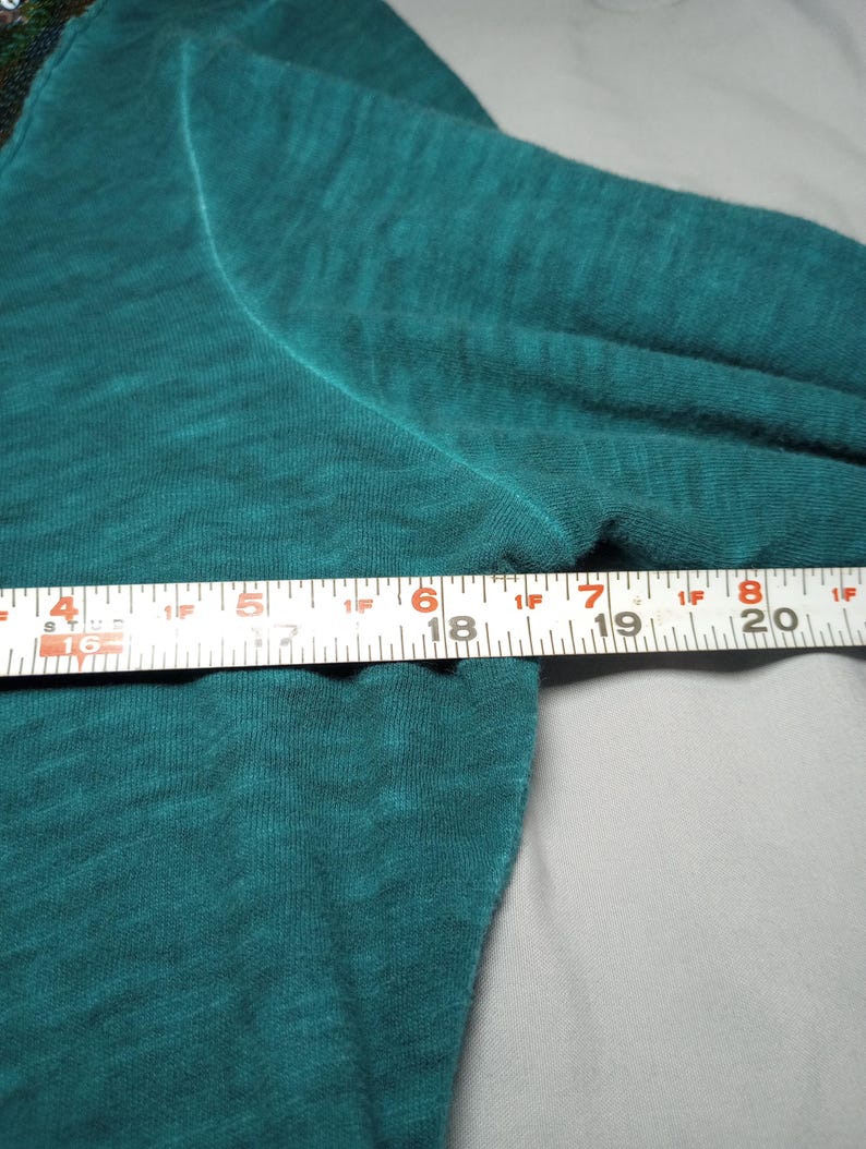 May include: Teal long-sleeve shirt with a crew neck. The shirt is made of a soft, textured fabric. A ruler is placed across the shirt, measuring approximately 6 inches.