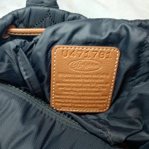 May include: Close-up of a navy blue quilted tote bag with a brown leather patch. The patch features the brand name "Brighton" and the number "U471781". The bag has a brown leather handle and a zipper closure.
